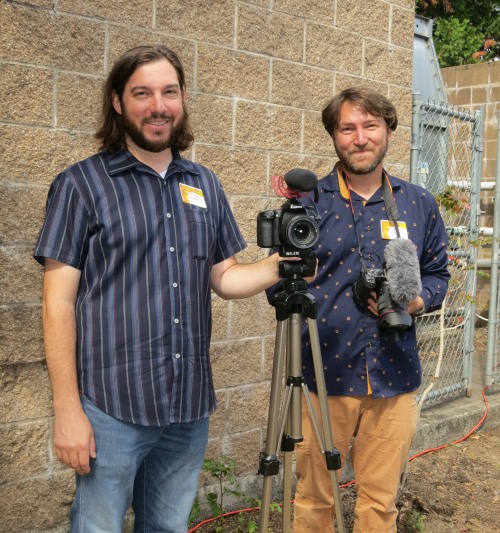 Two men stand in front of a concrete block wall holding camera equipment